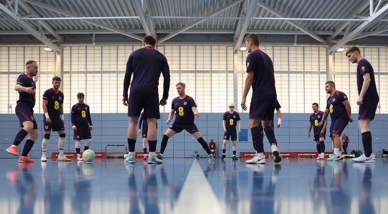 A photo of the team standing in an oval while training, all in dark blue football uniforms. Neil, on the far left of the image is in action- about to kick the ball as it comes towards him.