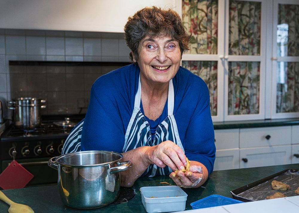 This portrait is of Penny, a woman in her late sixties with fair skin and short dark hair, in her kitchen leaning on her countertop smiling warmly at the camera while preparing dough with her hands - she is baking cookies. She wears an electric blue long sleeve top and a stripey navy and white apron. To her right there is a metal pot on the dark green countertop. To her left, a baking tray with cookie dough portions sits ready for the oven. Behind her, the traditional kitchen is decorated with floral-patterned curtains inside glass-front cabinets and a stove with shiny pots. The overall mood suggests warm, welcoming homely charm —– it feels like a snapshot of Penny happily sharing her cooking.
