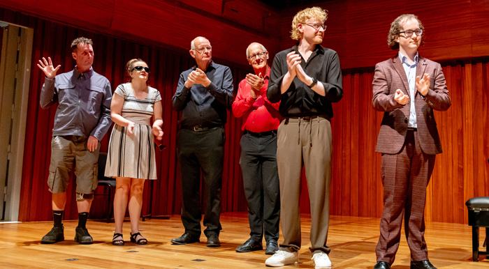 Six singers and musicians stand on a wooden stage in a warm-lit auditorium, clapping toward the audience. They are dressed in semi-formal clothing, arranged in a line, with wooden panelling behind them. The scene suggests they are acknowledging their performance.