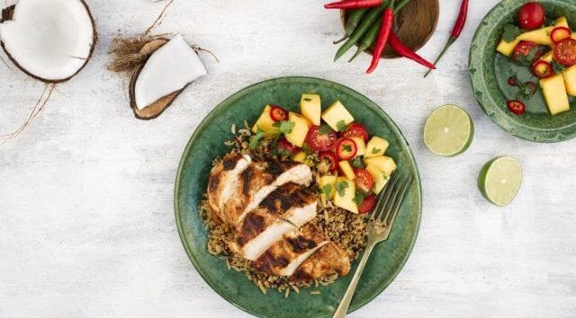 Birds eye view of a green plate in the middle of image, with chicken, rice and salad on the plate. The plate of food is surrounded coconut, peppers, lime and salad.