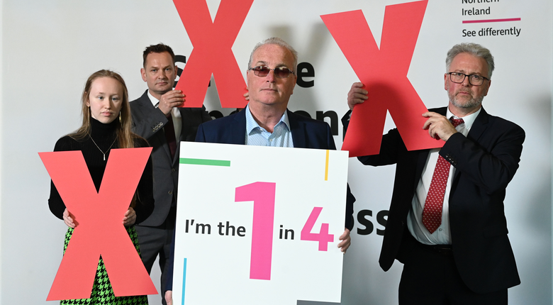 Four people standing in front of a white background with the RNIB Northern Ireland logo. One man is holding a sign which reads "I'm the one in four" as the other people hold up red crosses.