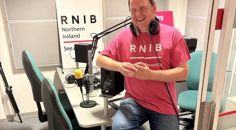 Joe perched on the studio desk looking relaxed arms crossed and laughing