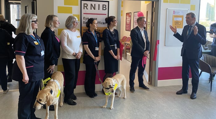 Staff from RNIB and South Tyneside and Sunderland NHS Foundation Trust outside the new ECLO room at Sunderland Specialist Eye Hospital. To the right is Ken Bremner the Trust's CEO. He is wearing a dark suit with his hands slightly raised as if to demonstrate the space. To his left is RNIB CEO Simon Antrobus wearing a dark suit and glasses and listening to Ken. To the left are members of staff including RNIB's two ECLOs, Mary Hall and Joe Hewitson. They are wearing the new ECLO uniforms which are navy blue and are both leading guide dogs. The entrance to the room is a tall white wall decorated with pink and yellow designs. Over the doorway is the sign: "Support for you and your vision starts here” in large black letters.