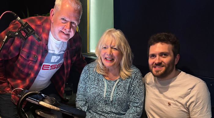 Cast members, left to right, Martin Trenamen, Alison Steadman and Reece Finnegansit, stand together in a recording studio, smiling toward the camera. A microphone and audio equipment are positioned in front of them. The person on the left wears a red plaid shirt, the person in the middle wears a blue patterned top, and the person on the right wears a light T‑shirt. The background shows a window and dim studio surroundings.
