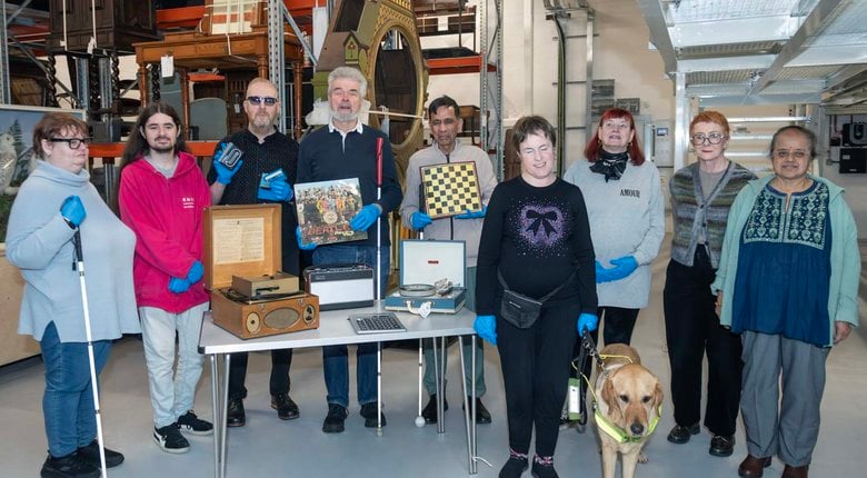 Nine members and a guide dog, stand smiling next to various items in the museum archives.