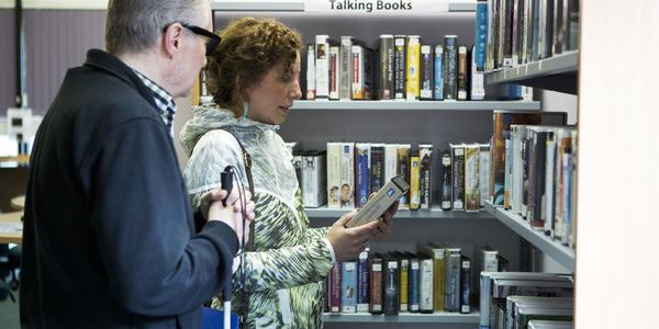 In a library setting, a man is to the left holding a cane, next to a woman who is holding a book, while a sign that says 'Talking Books' is above them in the background.