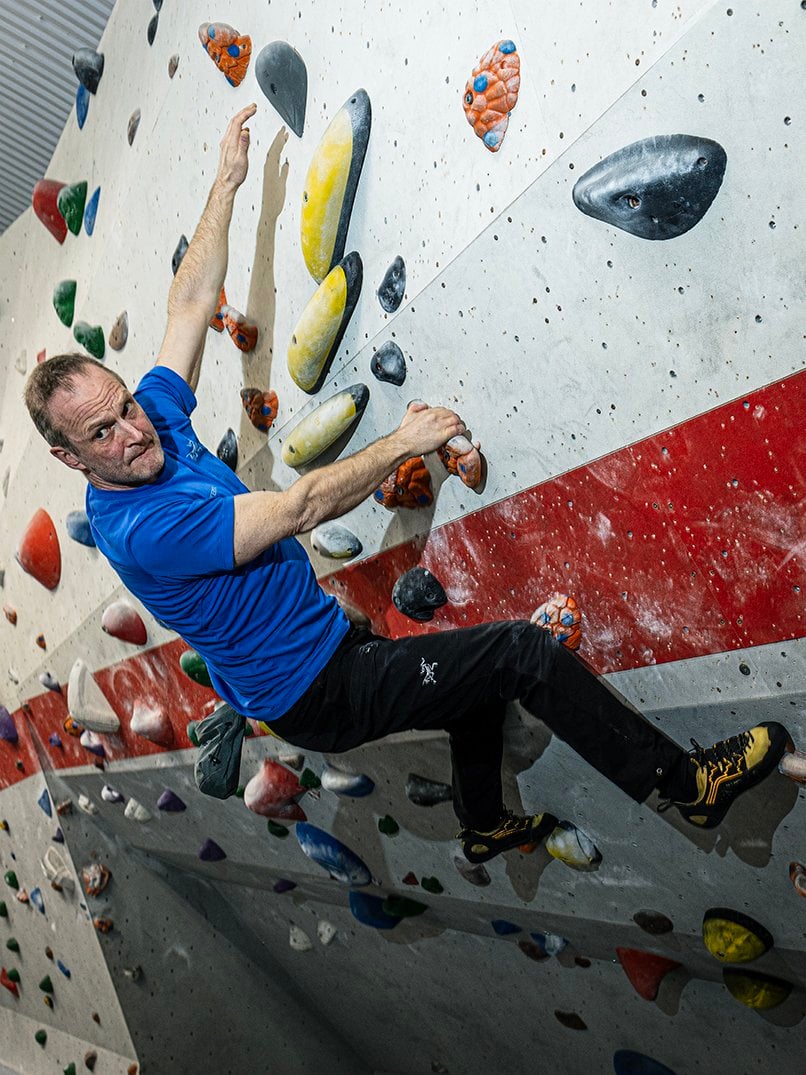 Red, a man in his fifties with fair skin and short dark hair, captured in a moment of intense concentration and physical effort. Red is mid-climb on an indoor bouldering wall, holding onto colourful climbing holds with both his hands and feet. His body is positioned diagonally, stretched across the wall, which emphasises the difficulty and athleticism involved in the climb. He’s wearing a bright blue t-shirt and black climbing trousers, along with specialised climbing shoes that offer grip and support. His left arm is extended upward, fingers reaching for the next hold, while his right hand is gripping an orange hold just below him. His face shows focus and determination, the tension in his muscles hinting at the challenge of the route.  The variety of climbing hold shapes, sizes and colours reflects the complicated technical nature of the route. This vibrant, high-energy image conveys Red’s strength, skill and mental precision involved in climbing.
