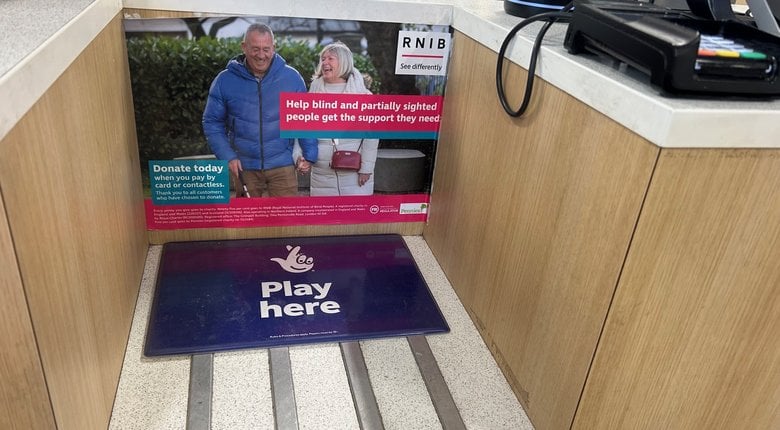 Image shows a poster on the counter of a petrol station. It shows a man holding a white cane and a woman. They are hand in hand and smiling. The poster text says 'Help blind and partially sighted people get the support they need - donate today'.
