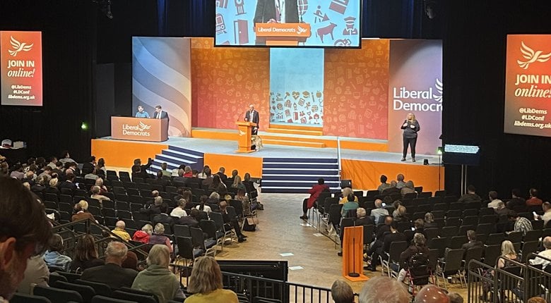 Steve Darling MP, accompanied by his guide dog Jennie, is stood behind an orange podium on the conference hall stage addressing a large crowd.