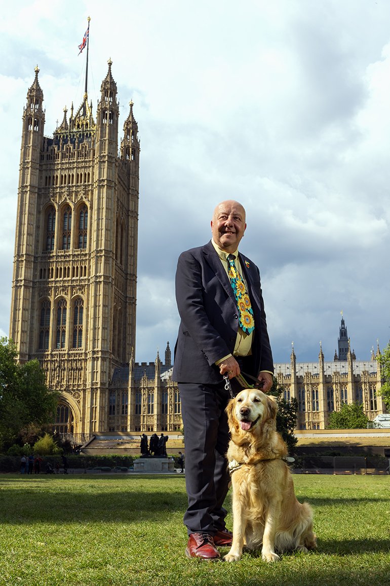 This portrait is of Steve, a man in his mid-fifties with fair skin and a bald head, facing towards the camera with his guide dog, Jennie, outside the Palace of Westminster in London. Steve stands on the grass, dressed in a dark suit with a light-yellow shirt and a bright cheerful sunflower-patterned tie, holding Jennie’s lead. Jennie, a golden retriever, sits calmly by his side. Behind them, the iconic Victoria Tower rises above the surrounding buildings under a partly cloudy sky with the Union Jack flying high at full mast, a nod to Steve’s work. The image conveys warmth, dignity and a strong sense of inclusion and civic pride.