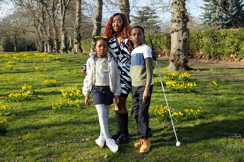 Sylvia, a woman in her mid to late thirties with black skin, standing with her two children in a sunlit Barking Park scattered with blooming yellow daffodils during springtime. Sylvia, holding her white cane, is positioned in the middle, and is wearing a stylish black-and-white patterned dress with lace up knee-high black boots. Her wavy long red hair and confident smile add vibrancy to the scene. Standing on her right is her daughter, who is about six years old with black skin and black hair, in a light jacket, navy skirt, and white tights, wearing white shoes and two yellow hair bows that complement her cheerful presence. On Sylvia’s left is her son, who is about ten years old with black skin and short black hair, dressed in a colour-blocked long-sleeve shirt, dark trousers, and tan boots. His right arm is gently resting on Sylvia, and the family’s closeness is clear in their body language and expressions.
They are standing in a serene park setting, framed by a line of leafless trees stretching into the background and surrounded by lush green grass dotted with early spring flowers. The light is golden, the mood is calm and proud, and the portrait speaks beautifully of strength, love, and family unity.
