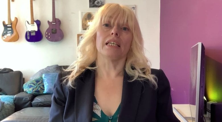 A white woman with blonde hair down to her shoulders, wearing a dark colour blazer talks to the camera.