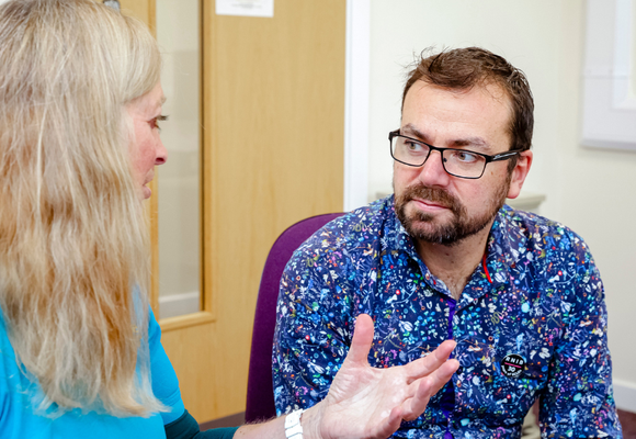 A white woman with blonde hair is talking to a man in an eye clinic and using her hands to gesticulate. The white man has brown hair, a beard and is wearing glasses and a bright blue printed shirt with a black RNIB pin badge on the front pocket.