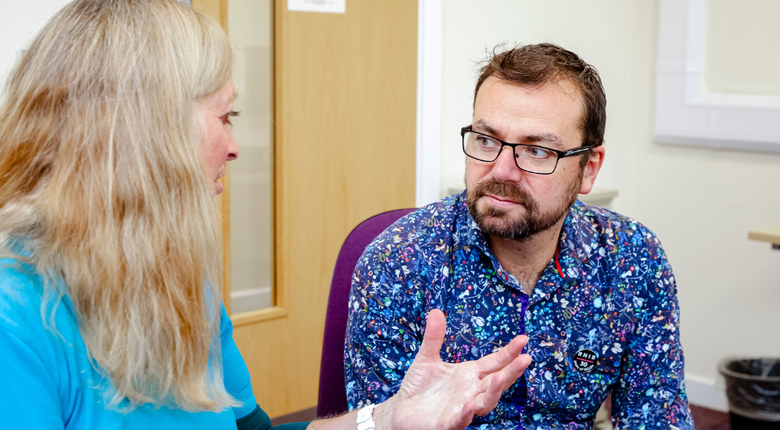 A white woman with blonde hair is talking to a man in an eye clinic and using her hands to gesticulate. The white man has brown hair, a beard and is wearing glasses and a bright blue printed shirt with a black RNIB pin badge on the front pocket.