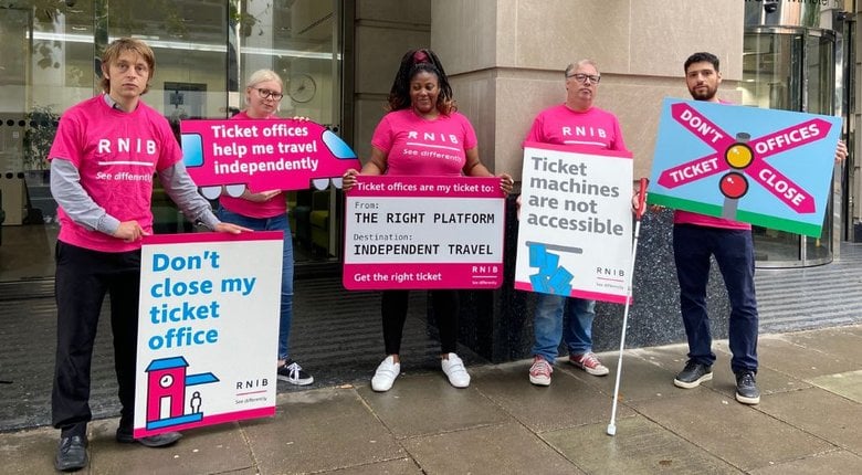 Five people wearing pink RNIB t-shirts and holding signs protesting railway stations ticket office closures, stand outside the Department for Transport.