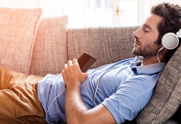 A man lying on a sofa, wearing headphones, listening to audio from an IPhone