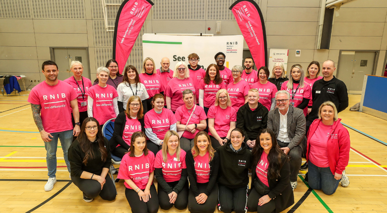 The RNIB Northern Ireland team. A group of around 30 people of varied ages. Almost all of them are wearing pink RNIB t-shirts. They are stood inside a large gym. Behind them are pink RNIB banners.