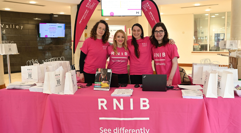 Four people wearing pink RNIB t-shirts stand behind a table in the entrance to the 2025 Tech Fair. The table is covered with a pink RNIB branded cloth and has lots of white paper bags on it also featuring RNIB branding.