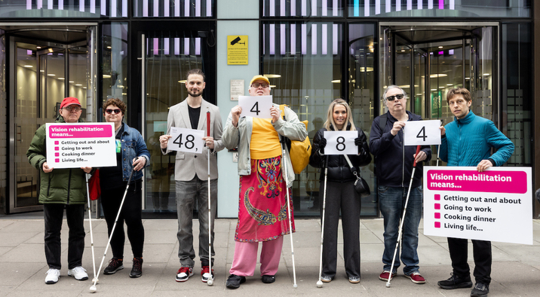 Seven staff and volunteers standing in a line in front of a glass building facade, the Department of Health and Social Care. They all have their long canes out in front of them on the pavement. The people in the middle of the group hold the numbers 48,484 on individual A4 sheets of paper, the number of people who signed the petition. The people on either end of the group hold placards that read "Vision rehab means: getting out and about, going to work, cooking dinner, living life."