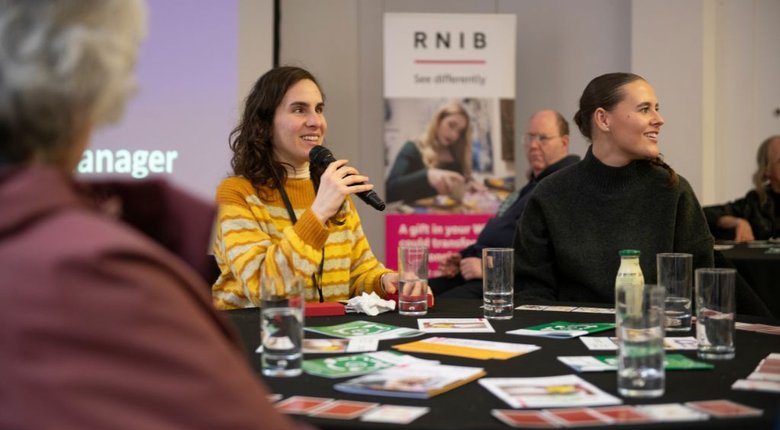 Alba and other guests are sat around a circular table covered in leaflets and glasses of water. Alba is holding a microphone and is wearing a yellow striped jumper and has long brown hair. In the background there is RNIB signage and other guests are listening to Alba speak.