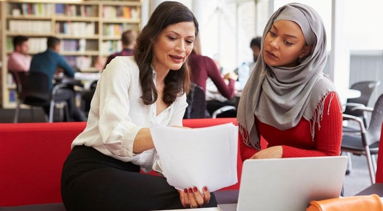 Two women sat side by side on a red sofa are reading a document together, they also have a laptop open in front of them. The woman on the right is wearing a hijab.