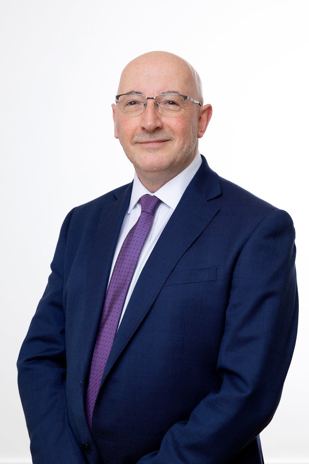 Jim Harra, a white man in his fifties wearing glasses and a navy blue suit standing professionally and smiling at the camera against a white background