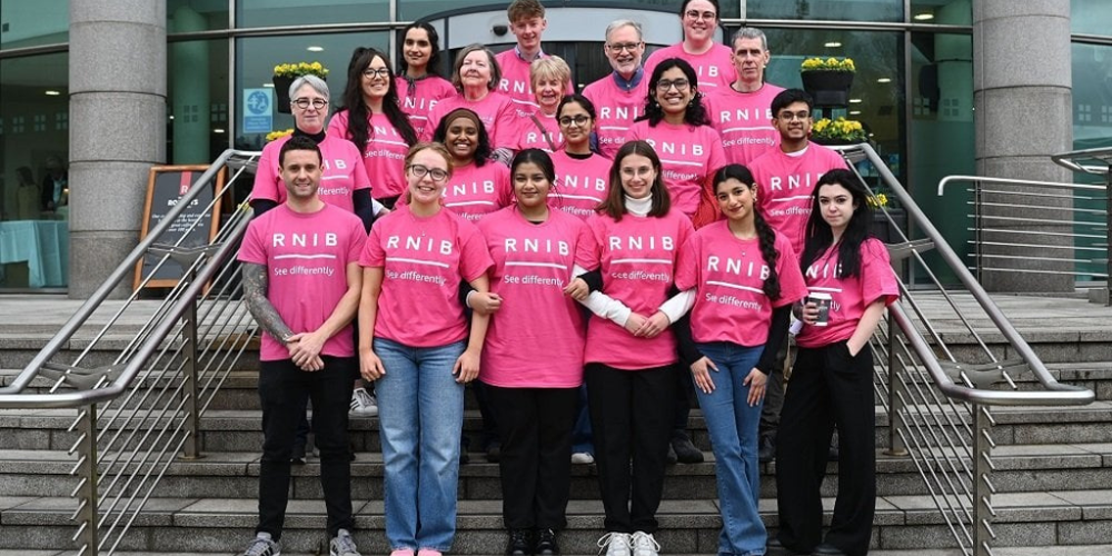 Image description: a line of 19 volunteers on steps outside a modern glass building, wearing RNIB pink t-shirts.