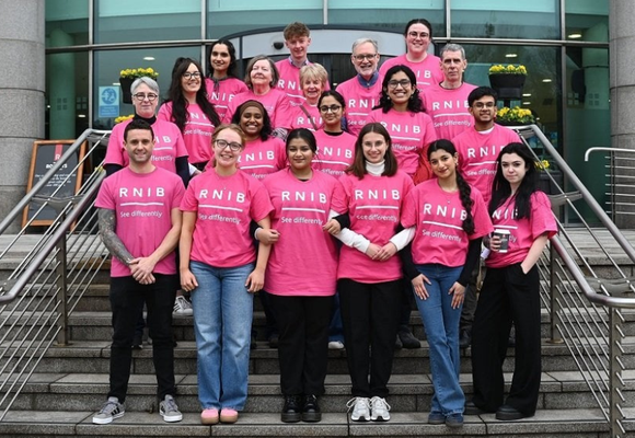 Image description: a line of 19 volunteers on steps outside a modern glass building, wearing RNIB pink t-shirts.