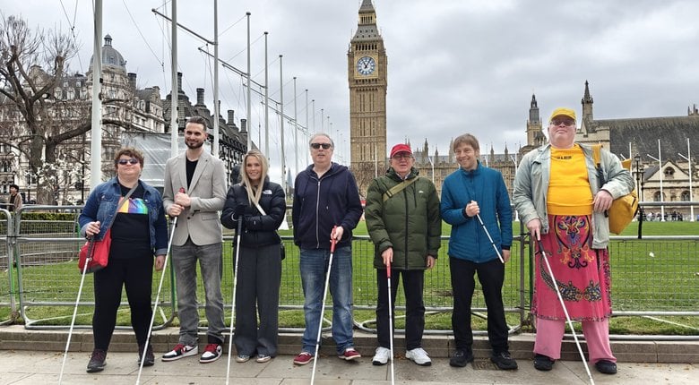 Seven staff and volunteers standing in a line at Parliament Square. They all have their long canes out in front of them on the pavement. Behind them, a grey, overcast sky is broken only by the solitary tower of Big Ben in the background.