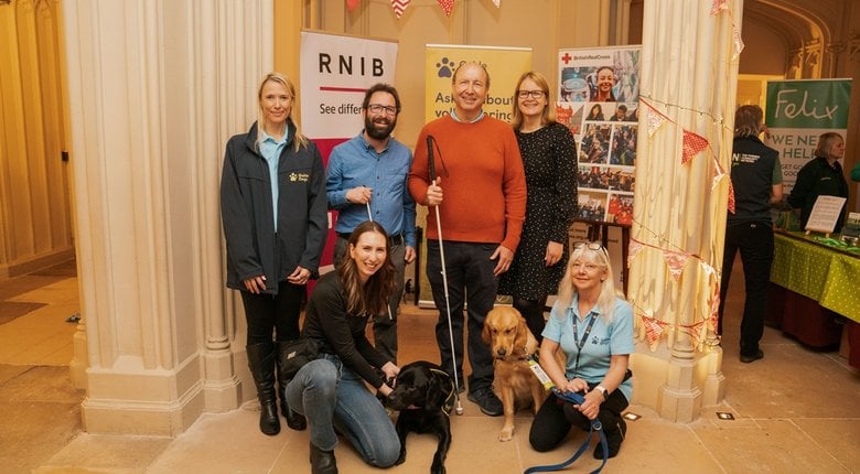 RNIB volunteers Simon Holt and Jonathan Abro are stood in the centre of the photograph. Jonathan is wearing an orange sweater and Simon has a blue shirt on, they are both holding canes. Also pictured is RNIB's Jo Keller who is stood next to Jonathan as well as a representative from Guide Dogs and two guide dog handlers with their dogs are also crouching on the ground.