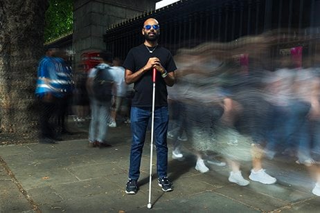This portrait is of Yahya, a man in his early thirties with brown skin, a shaved head and a neatly trimmed black beard, standing confidently and centred amidst a bustling crowd on a London pavement. He holds a white cane and wears dark sunglasses, a black t-shirt, and blue jeans. The motion blur of people rushing past him emphasises both the fast pace of the world and Yahya’s stillness, presence, and resilience within it. The visual contrast between Yahya’s calm composure and the surrounding blur powerfully symbolises strength, independence and the often-overlooked experience of navigating the world without sight.