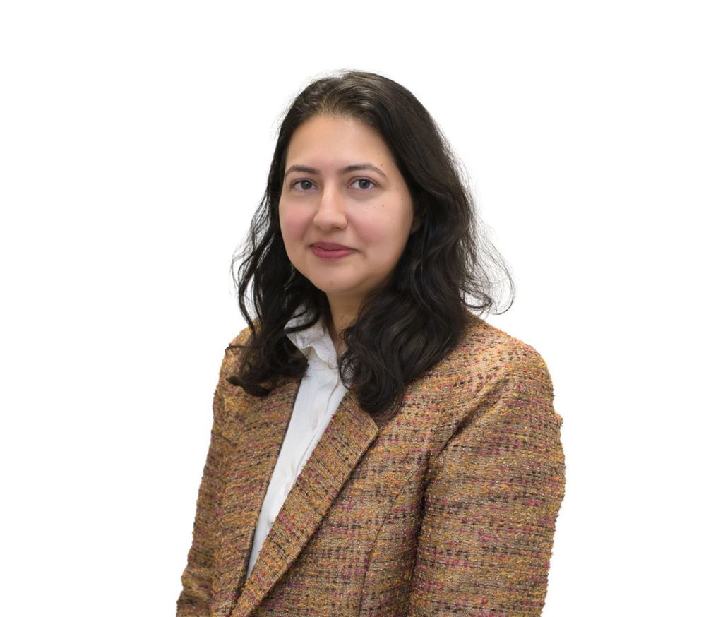 A picture of Zayna Ali, a woman with black hair wearing a brown blazer and white shirt smiling at the camera