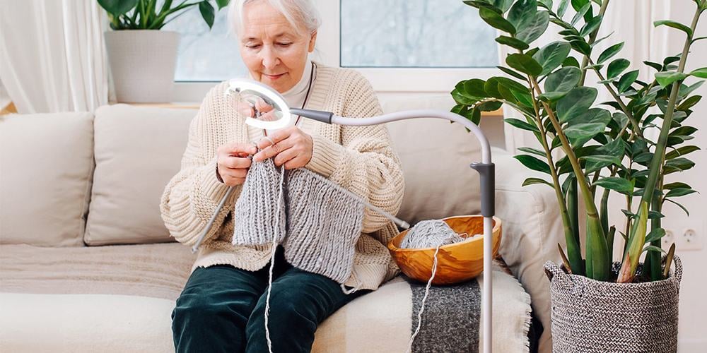 An elderly woman with white hair wearing a cream cardigan and bottle green trousers is sat down on a sofa knitting with light over the thread and knitting needle.