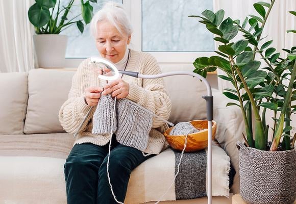 An elderly woman with white hair wearing a cream cardigan and bottle green trousers is sat down on a sofa knitting with light over the thread and knitting needle.