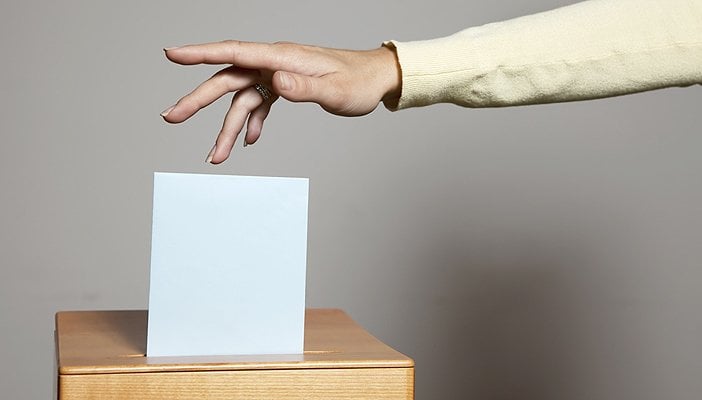 A hand dropping a folded ballot into a ballot box.