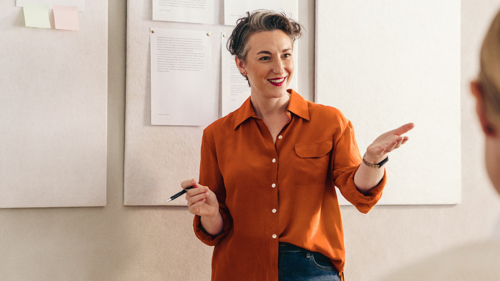 A woman in an orange shirt with short hair, standing and engaging with a room of people.