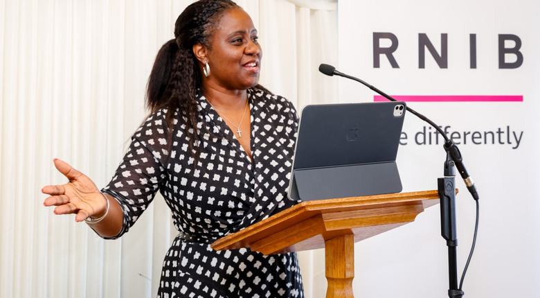Marsha de Cordova MP speaking at the launch of the RNIB training which highlights the importance of emotional support at the point of a sight loss diagnosis. Marsha is wearing a black and white dress and standing in front of a white backdrop and an RNIB logo.