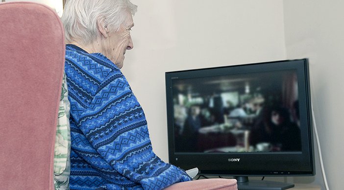 An older person sat in a light pink armchair watching TV.