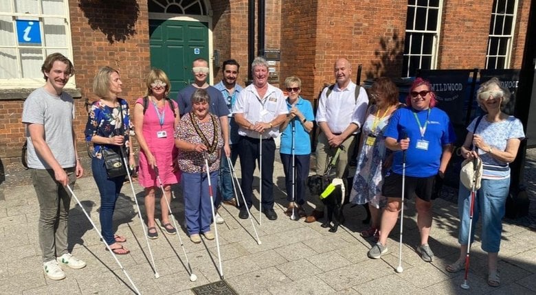 Steve in the middle of a large group of people on a sunny day in front of a red brick building. Everyone is holding a long cane out in front of them and smiling.